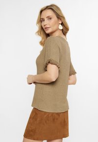 Khaki pletený svetr s texturovaným povrchem a třásněmi na krátkých rukávech, doplněný hnědou semišovou minisukní. Minimalistický design a měkká látka.