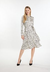 Beige mekko, jossa on musta ruutukuvio, pitkät hihat, vyö vyötäröllä ja levenevä helma. Yhdistetty mustiin korkokenkiin.
