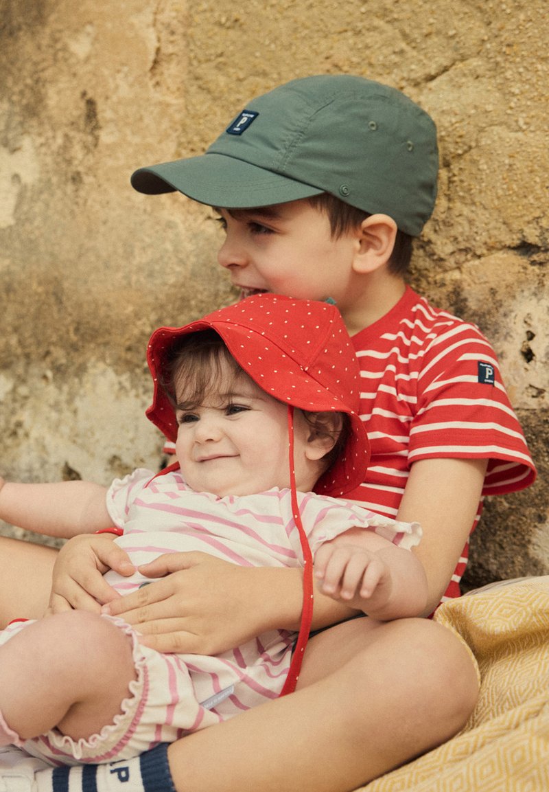 Punainen ämpärilakki, jossa on valkoisia pilkkuja, lapsen päässä, pukeutuneena vaaleanpunaiseen ja valkoiseen raidoitettuun asuun, istuu vieressä sisaruksen kanssa, joka pitää vihreää lakkiä ja raidoitettua paitaa.