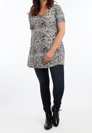 Vrouw draagt een grijze tuniek met korte mouwen en slangenprint, een kruiselings gevormde halslijn, gecombineerd met donkere strakke jeans en zwarte veterlaarzen.