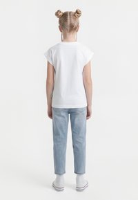 Copil cu părul blond aranjat în două cocuri, purtând un tricou alb simplu, blugi bleu deschis, șosete albe și adidași albi, stând cu fața spre spate.