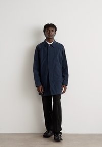 Mornarsko plavi kaput s okovratnikom, zatvaračem na dugmad, dugim rukavima i ravnim krojem, uparen s crnim hlačama i cipelama.