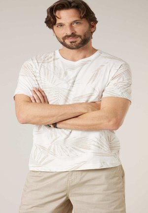 Man met baard en golvend bruin haar staat met gekruiste armen, draagt een wit T-shirt met beige bladpatronen en beige broek.