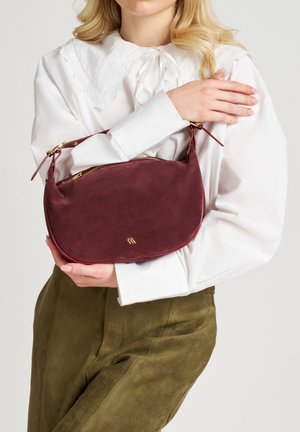 Sac à main en suède bordeaux avec une forme courbée, des garnitures dorées et une fermeture éclair. Le sac dispose d'une courte bandoulière pour un port à la main ou à l'épaule.
