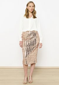 Balta ilga rankovė bluzonė derinama su kelių ilgio, rožinio aukso spalvos blizgučiais padengta sijonu. Sijonas turi vertikalią blizgesio raštą ir subtilią tekstūrą.