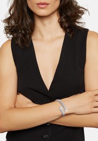 Pulsera trenzada gris con un cierre hexagonal plateado, llevada en una muñeca desnuda sobre una blusa sin mangas negra, mostrando una textura suave.