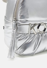 Mochila plateada metálica con textura suave, cuerpo plisado y herrajes plateados. Presenta un acento con el logotipo y cierre de cremallera.