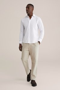 Balti ilgomis rankovėmis marškiniai, susegti, prigludusio kirpimo. Šviesiai smėlio spalvos kelnės, tiesaus kirpimo. Tamsiai rudi batai su faktūruotu paviršiumi. Neutralus fonas.