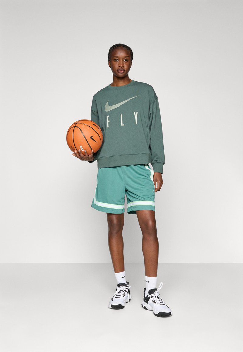 Felpa verde con il testo "FLY" e il logo Nike, abbinata a pantaloni corti color turchese con strisce bianche. Tiene un pallone da basket e indossa scarpe nere e bianche.