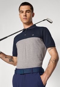 Marineblå og grå kortermet polo skjorte med krage, tre knapper og CK-logo; kombinert med et blått strukturert belte og marineblå bukser. Golfkølle i hånden.