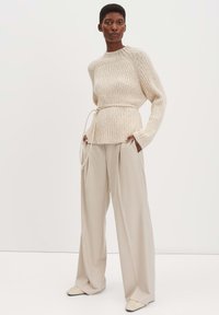 Beige ribbeltrui met een taille met riem en lange mouwen, gecombineerd met lichtbruine wijde broek en beige instapper schoenen.