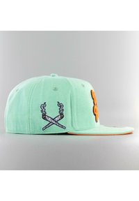 Vihreä mintunvärinen hattu, jossa on tasainen lippa, oransseilla yksityiskohdilla, ja vasemmalla puolella brodeerattu ristiin aseteltu esine, jossa on savuyksityiskohtia.