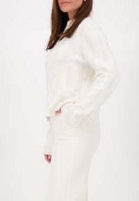 Wit gebreid sweater met groot subtiel logo patroon, ronde halslijn en lange mouwen; gecombineerd met strakke crème kleurige pantalons.