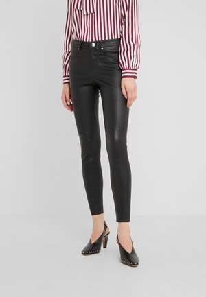 Kobieta ubrana w czarne dopasowane skórzane spodnie, czarne sandały na wysokim obcasie z odkrytą piętą oraz długą, białą bluzkę z czerwonymi pionowymi paskami.