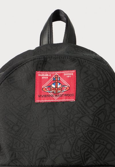 Melns audums mugursoma ar teksturētu virsmu, kurai ir sarkans uzlīme, kurā rakstīts "Vivienne Westwood", un sarežģīti raksti materiālā.