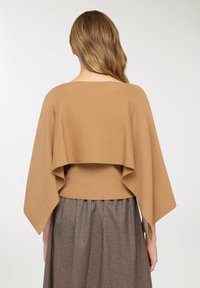 Maglione a mantella lavorato a maglia in cammello con un design fluente e drappeggiato sul retro, abbinato a una gonna plissettata marrone a quadri.