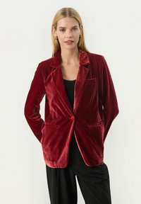 Blazer rosso vellutato con un bottone singolo, rever a scatto, due tasche frontali e una leggero lucido. Indossato sopra una maglietta nera, abbinato a pantaloni neri.