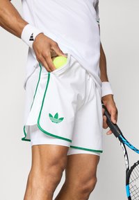 Shorts de tennis blancs avec des bordures vertes et un logo Adidas, comportant une poche latérale tenant une balle de tennis jaune. Raquette de tennis noire en main.