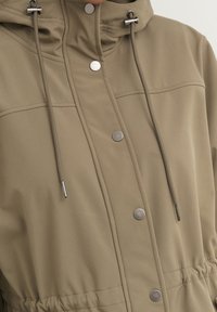 Giacca khaki con cappuccio realizzata in tessuto morbido, caratterizzata da una chiusura con coulisse, bottoni a pressione argentati e dettagli arricciati in vita per un look aderente.