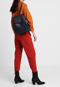 Zaino in pelle blu navy con un design strutturato, pattina frontale e un accento di cerniera rossa. Il modello indossa una camicia color ruggine e pantaloni rossi, con stivaletti neri con tacco.