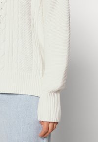 Witte kabelgebreide trui met gestructureerde patronen, geribbelde manchetten en zoom, en een ontspannen pasvorm, gecombineerd met lichtblauwe spijkerbroek.