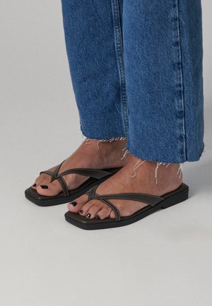 Piedi con smalto nero che indossano infradito nere con punta quadrata e jeans blu con orlo sfrangiato su uno sfondo chiaro uniforme.