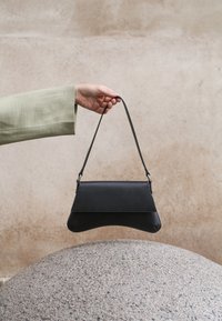 Χέρι που κρατά μια μαύρη δερμάτινη τσάντα χειρός από το λουρί της πάνω από μια υφή από πέτρινη επιφάνεια με ουδέτερο μπεζ φόντο.