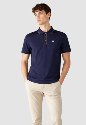 Uomo con polo blu navy con rifiniture beige e bottoni, abbinata a pantaloni beige chiaro, in piedi con le mani in tasca, guarda di lato.