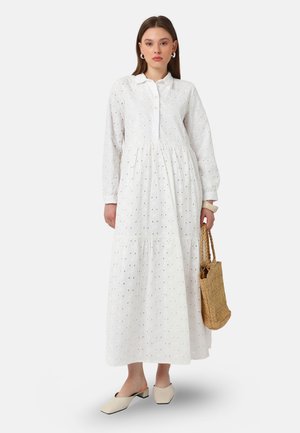 Mujer con un vestido largo blanco con ojales y mangas largas, sosteniendo un bolso de paja tejido y llevando zapatos beige sin cordones.
