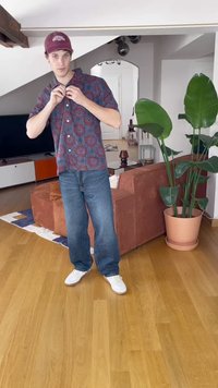 Rövid ujjú ing piros és kék körmintával, laza kék farmernadrággal és fehér sportcipővel párosítva. Belső tér növényekkel.