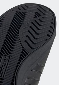 Semelle de chaussure Adidas noire avec un motif de bande de roulement détaillé et le logo Adidas près du talon, montrant les caractéristiques de texture et d'adhérence.