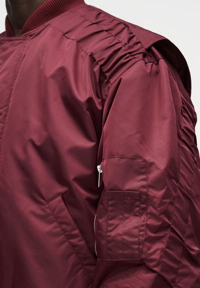 Bordová bomber bunda z hladkého nylonu, s nariasenou látkou na pleciach, zipsovou kapsou na rukáve a rebrovaným golierom.