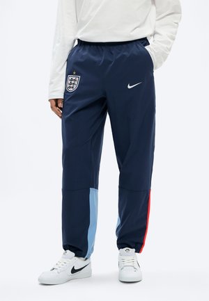 Personne portant un pantalon de jogging Nike de l'équipe d'Angleterre bleu marine avec des panneaux de jambe bleu clair et rouge, ainsi que des baskets blanches Nike.