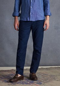 Chino blu navy in cotone misto, con vestibilità slim e frontale piatto. Abbinati a mocassini marroni, su uno sfondo di un tappeto testurizzato.