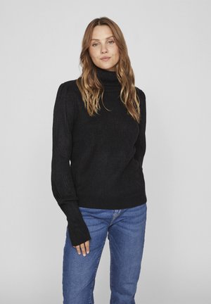 Pull noir en maille à col roulé avec texture côtelée. Présente des manches longues et des poignets ajustés, associé à un jean bleu pour un look décontracté.