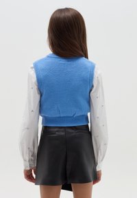 Blau gestrickte Weste mit geripptem Saum, getragen über einem weißen Langarmshirt mit Herzmuster, kombiniert mit schwarzen Leder-Shorts.