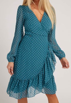 Robe portefeuille bleu sarcelle à pois blancs, manches longues transparentes, ourlet à volants et taille cintrée sur une personne debout aux longs cheveux blonds.