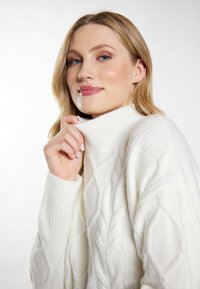 Biały, dzianinowy sweter z wysokim kołnierzem i teksturowanym wzorem warkocza. Ręka modelki delikatnie ciągnie za kołnierz. Gładki, miękki wygląd.