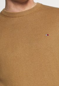 Maglione marrone a maglia con scollo rotondo, con un piccolo logo sul lato sinistro del petto. Texture liscia e polsini a coste.
