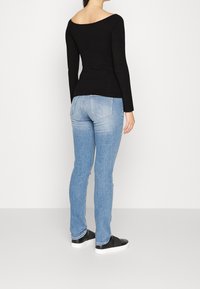 Frau, die mit dem Rücken zur Kamera steht, trägt ein schwarzes langärmeliges Oberteil, helle blaue schmal geschnittene Jeans und schwarze Slipper mit weißen Sohlen.