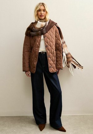 Braune gesteppte Jacke mit großen Knöpfen, kombiniert mit hoch taillierten dunklen Jeans aus Denim. Ein mehrfarbiger Schal liegt locker über der Jacke.