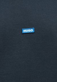 Μπλε ορθογώνιο ετικέτα με λευκό κείμενο "HUGO" σε σκουρόχρωμο γκρι φόντο υφάσματος, κεντραρισμένο στο κάδρο.