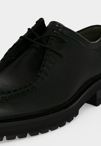 Chaussure habillée en cuir noir avec couture en relief autour de l’orteil, fermeture à lacets et semelle texturée robuste.