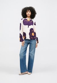 Krém színű, virágos pliszírozott blúz lila és narancssárga virágokkal; széles szárú kék farmernadrággal és világos színű balerinacipővel párosítva.