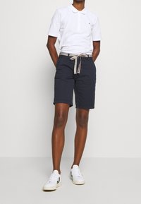 Vit polo med tre knappar, tillsammans med marinblå shorts med en midja som har ett vävt bälte. Vita sneakers kompletterar outfiten.