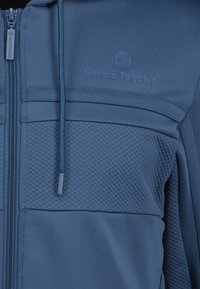 Felpa con zip blu, pannello frontale texturizzato, tessuto del corpo liscio e cappuccio regolabile. Presenta dettagli del logo discreti sul petto.