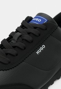 Μαύρο δερμάτινο αθλητικό παπούτσι με υφασμένη επάνω επιφάνεια, στρογγυλή μύτη και αντιθετική μπλε ετικέτα· διαθέτει μαύρα κορδόνια και λογότυπο στο πλάι.