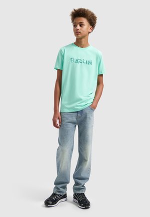 Tienerjongen met krullend haar, een mintgroen T-shirt met "Berlin" opdruk, lichtblauwe spijkerbroek en zwarte sneakers, staand met één hand in de zak.