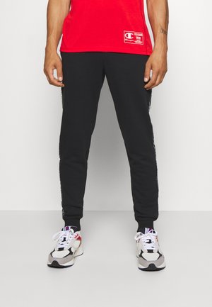Mand iført en rød T-shirt, sorte joggerbukser og hvide sneakers med sorte og røde detaljer, stående mod en ensfarvet baggrund.
