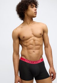 Male model with curly hair wearing black Tommy Hilfiger boxer briefs with red waistband, looking to the side against a plain background.